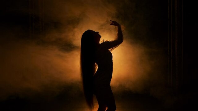 Side view silhouette of young woman dancing in dark smoky studio against background of yellow lights. Female with long hair performs sensual movements with hands touching body. Close up. Slow motion.