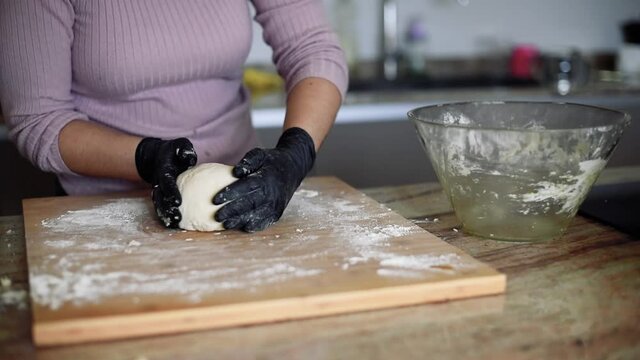 Una mujer en la cocina amasando una masa de pizza en una tabla de madera con harina mientras usa guantes negros.