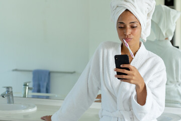 Mixed race woman wearing bathrobe brushing teeth and using smartphone in bathroom