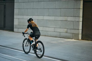 Tuinposter Rear view of professional female cyclist in black cycling garment and protective gear riding bicycle in city, passing buildings while training outdoors on a daytime © Friends Stock