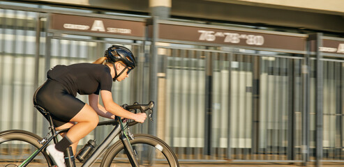 Professional female cyclist in black cycling garment and protective gear riding bicycle in city center rushing and passing buildings