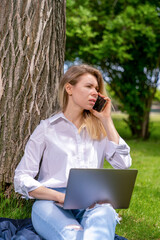 a girl sits on the grass by a tree in a country park with an open laptop on her knees and communicates on a mobile phone