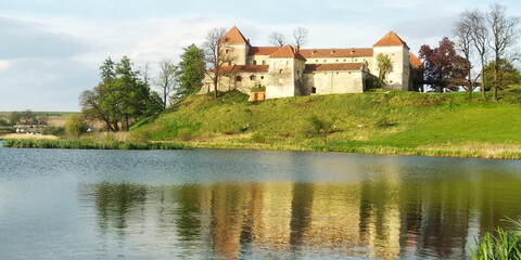 Fototapeta na wymiar Amazing panorama of Svirzh medieval castle, reflected in water of nearby lake under high blue sky with clouds