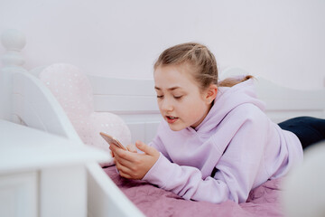 A Caucasian teenage girl uses a smartphone while resting on a bed in her room. The girl is video chatting with friends from school. Isolation. Remote learning. Coronavirus