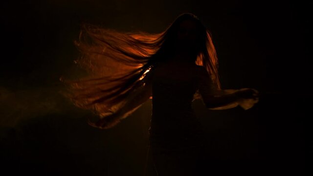 Young beautiful woman whirls and waves her hair in dark studio against the backdrop of bright soffit with yellow light. The female is posing and gracefully moving her arms in slow motion. Silhouette.