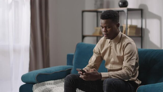 young afro-american man is using smartphone sitting on couch in apartment, male internet user, viewing social nets