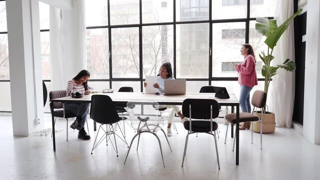 In the office. Three businesswomen work together without masks, keeping a safe distance. Two of them are working at the desk and one is chatting on her smart phone near the window.