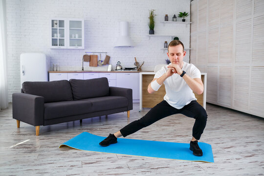 A strong athletic man in a T-shirt does exercises in his spacious apartment with a minimalistic interior. Healthy lifestyle during the quarantine period. Fitness at home
