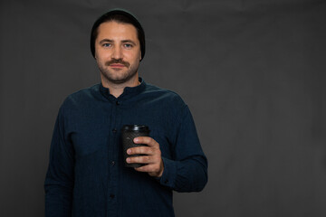 Handsome unshaven guy in knitted cap posing with disposable cup looking at camera on gray studio backdrop copy space. Morning wake-up coffee, take-away coffee, coffee shop or snack bar advertising