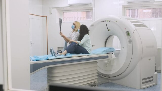 Woman doctor radiologist explains the results of CT scanning for young female patient, showing the snapshot with images, observing and analyzing CT scan in modern clinic beside modern CT scanner