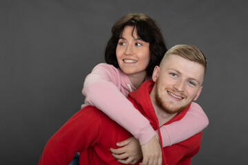young happy man carrying brunette girlfriend on his back. Cheerful vivid casual dressed couple fool around. Happy couple having fun piggyback ride in love on gray studio backdrop. Prenuptial agreement