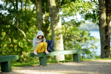 Pretty teenage girl using a smart phone sitting on a bench. Cute young girl having fun during forest hike on beautiful summer day. Teenagers addiction to new technology.