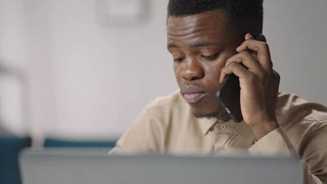 black man is communicating by mobile phone, sitting at home and working with laptop, closeup portrait of afro-american
