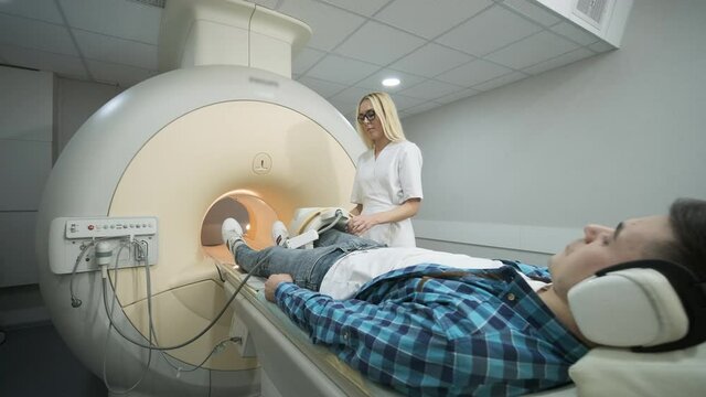 Woman doctor makes knee-joint MRI scanning. Young man patient on automatic table enters into a closed-type mri machine using noise isolation headphones. Modern equipment, coil on the patient's knee