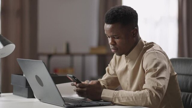 black male office worker or freelancer is using smartphone during work with laptop, sitting at table, surfing internet