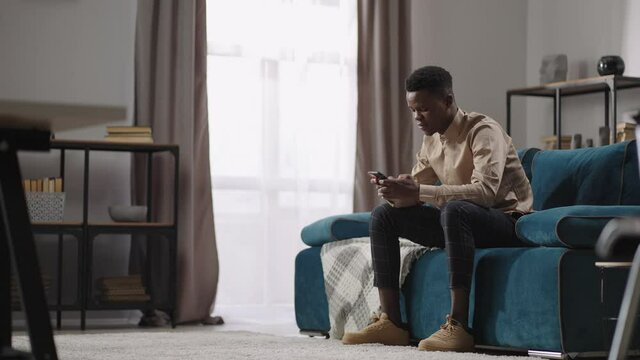 afro-american man is using smartphone for communication and playing games, sitting at home alone, black guy is typing message and scrolling screen