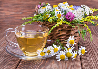 Herbal tea with the chamomile flowers in a glass cup and fresh wild flowers in a basket on a wooden background.