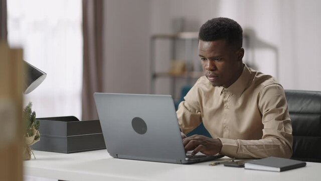 young black man is working with laptop at home, sitting at table and typing, male student or freelance worker, medium portrait of working guy