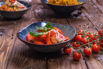 Sliced tomatoes with onions in a plate on a brown wooden background, decorated with cherry tomatoes, coarse salt and pepper peas.