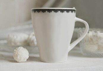 White cup with tea, coconut candy balls, marshmallows on a plate on a wooden background.