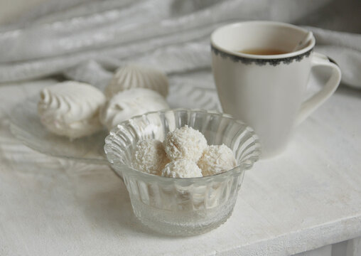 White cup with tea, coconut candy balls, marshmallows on a plate on a wooden background.