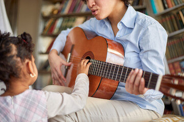 A cute little girl is so interested in her mother guitar playing while spending time at home together. Family, home, playtime