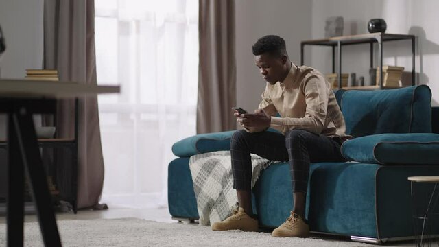 young black man is playing video game in smartphone, sitting at home alone, afro-american guy is communicating in social nets in mobile phone