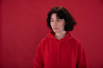 Fototapeta na wymiar tired brunette Girl in red hoodie posing on red studio backdrop copy space for ads text. Pain, fatigue, tension concept. Loneliness, desire to talk to someone. Psychotherapist, psychologist ads