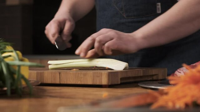 chef in restaurant kitchen cuts celery with sharp, shiny knife. man prepares food for family and cuts celery on wooden cutting board, vegetarian food salad.
