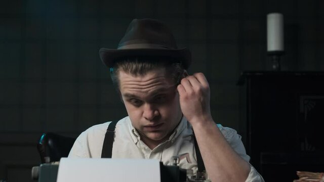 Detective or gangster or writer in white shirt and suspenders sits at table and prints something on typewriter. puts a cigarette behind the ear.