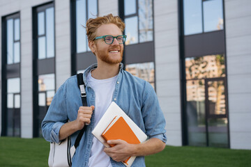 Smiling university student with backpack and book walking in university campus, education concept. Portrait of happy handsome man wearing stylish eyeglasses looking away on the street, copy space 