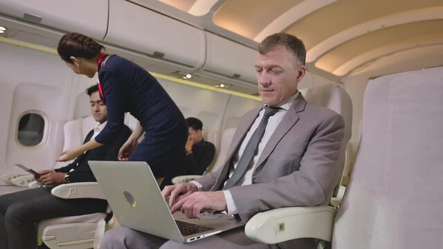 Senior professional businessman using computer laptop on airplane. Happy handsome business man in suit sitting and smiling near window with typing on keyboard while using laptop on first class flight 