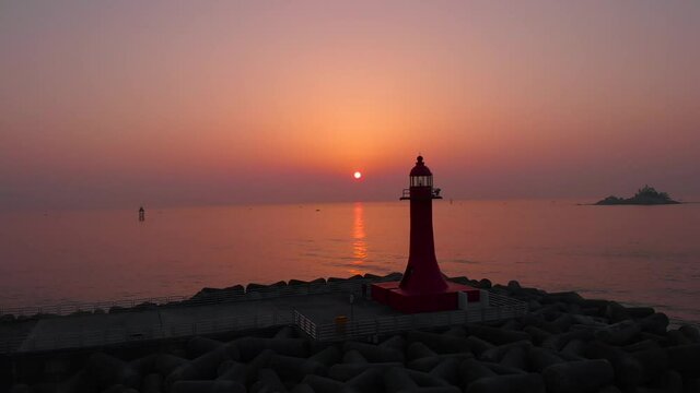 Sunrise at the east ocean with red glow sky. Red lighthouse stands on wave breaker at sea. Beautiful nature moment took with drone. 4K  