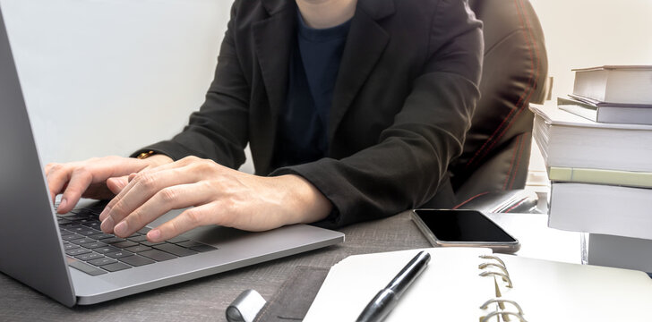 Casual Businessman Typing Laptop With Pile Of Books And Personal Blank Diary On Wood Table, Natural Light. Asian Executive Manager In Semi-formal Suit Working At The Office. Management Concept