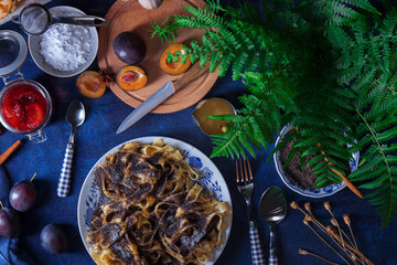 Sweet pasta dessert, noodles with poppy seeds, plum compote from fresh plums, dark background, blue table decorated with fresh flowers. Dry whole poppy plant in background.