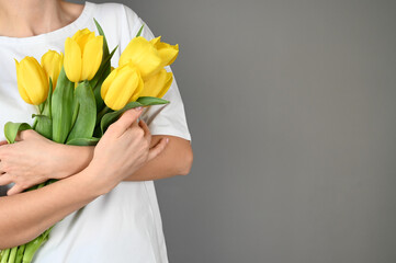 Cropped view of woman in white t-shirt holding yellow tulips against a gray wall with copy space.