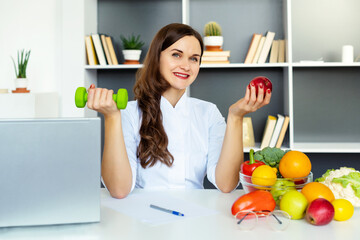 Smiling Female nutritionist holding a red apple and dumbbell showing healthy vegetables and fruits.