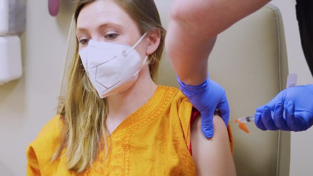 COVID 19 Vaccine Shot in the Arm of Young Adult Female Woman Wearing a Medical Mask, Corona Virus Injection Administered by Nurse in Shoulder in Doctor’s Office with the Patient Person’s Face Visible