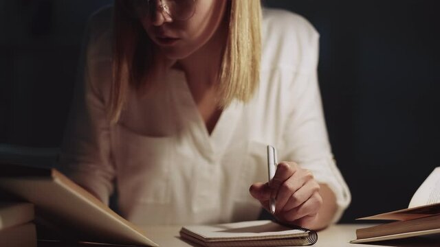 Late study. Academic research. Knowledge science. Intelligent smart left-handed female student in eyeglasses working from home on project writing essay with textbooks late at night at dark workplace.
