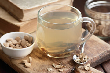 Burdock root tea in a glass cup with homeopathic pills, herb and old apothecary books nearby on wooden rustic background, closeup, naturopathy, natural medicine and homeopathy concept