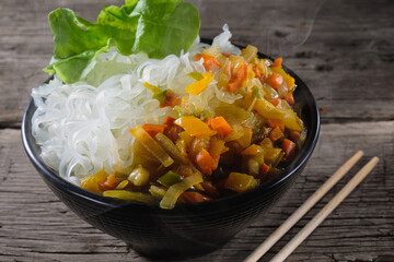 A plate of rice noodles stands on an old wooden table top. The side dish consists of fried onions, carrots, paprika.