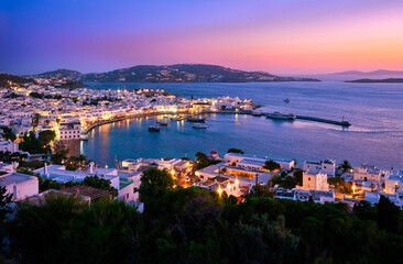 Fototapeta na wymiar Colorful view after sunset over town of Mykonos, Cyclades, Greece, harbor and port, cruises, ship, whitewashed houses. Colorful town lights up.