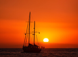Fototapeta premium Silhouette of sailing boat with sails down against sun at sunset, sun glare on sea waters. Romantic seascape. Sailboat go to sea