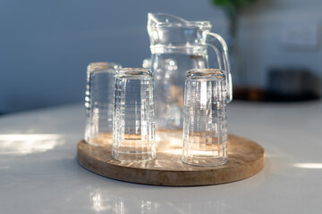 Glass water bottle jar with water glasses inside house lit by natural light from window on kitchen bench