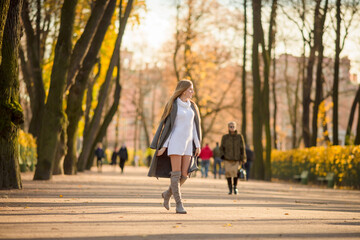 Happy young woman walks in the autumn park. An attractive woman with light brown hair and a gray coat is walking down the alley. Beautiful autumn mood. Fall season.