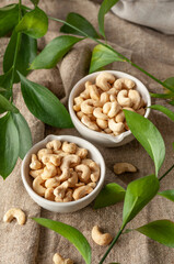 Cashew nuts in two white cups. Fabric background. Nearby cashews and green leaves. View from above.