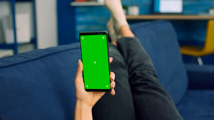Freelancer woman lying on sofa looking at mock up green screen chroma key smartphone in vertical mode. Caucasian female browsing on internet using isolated phone for social media network