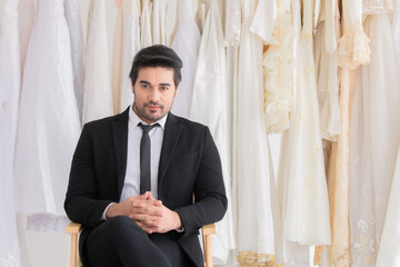 Handsome mix race beard groom hold flowers bouquet waiting for bride while fitting wedding dress,. happy smart man with tuxedo sitting in fitting room, man smiling look to the window with copy space