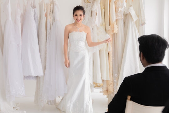 Happy mix race wedding concept, Caucasian groom with beard  sitting to help bride fitting choosing wedding dress in studio, happy beautiful Asian girl show her lace wedding dress to her man