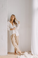 Young blonde woman playing with her hair, wearing a white shirt and pants, indoors, on white wall background.